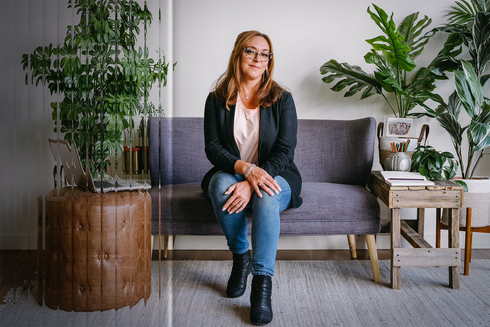 Elizabeth Miner sits on a sofa surrounded by drawing materials, a laptop, and potted plants. A clear reeded glass texture overlays part of the image.