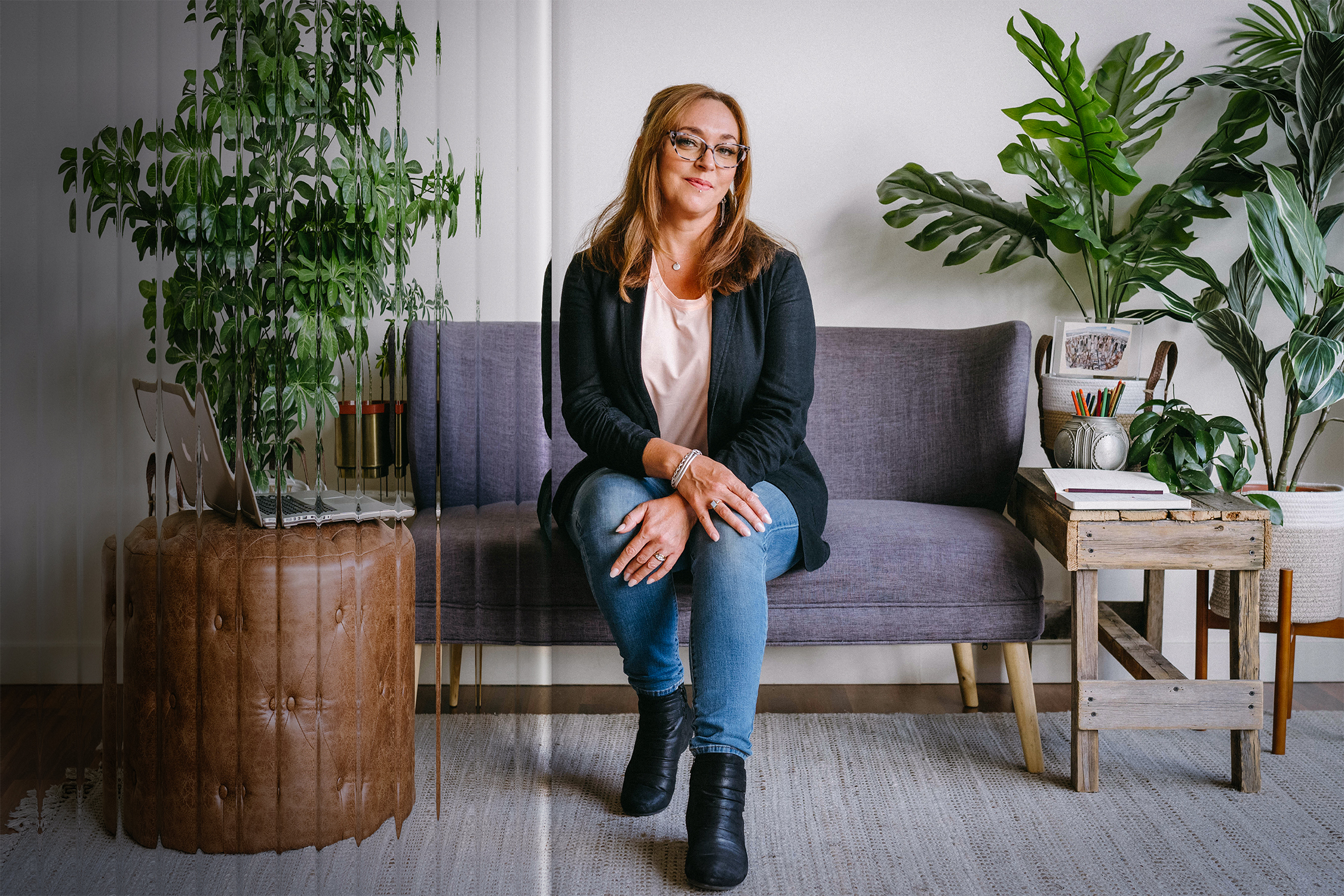 Elizabeth Miner sits on a sofa surrounded by drawing materials, a laptop, and potted plants. A clear reeded glass texture overlays part of the image.